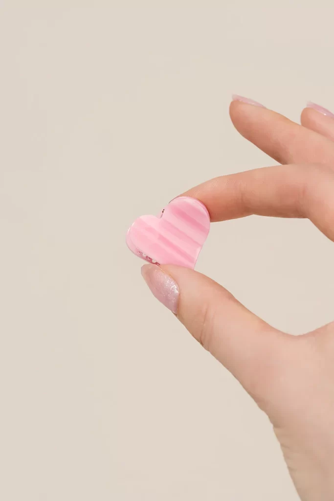 A delicate woman's hand is holding a tiny heart-shaped pink PixieWoo claw clip.