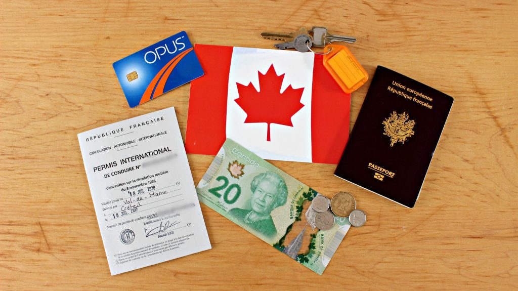 L'image d'une surface en bois sur laquelle est déposé un passeport français; un billet de 20$ et des pièces de monnaies canadiens; un permis de conduire, une carte opus, des clés et un drapeau du canada