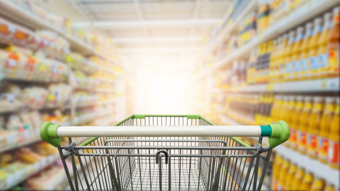 Image d'un chariot de magasinage vide au milieu d'un rayon d'épicerie les étagers contiennent des articles sous forme de bouteilles, de sacs en plastiques et des boites en cartons pas très bien apparentent car l'image est flouté vers un fond bien illuminé.