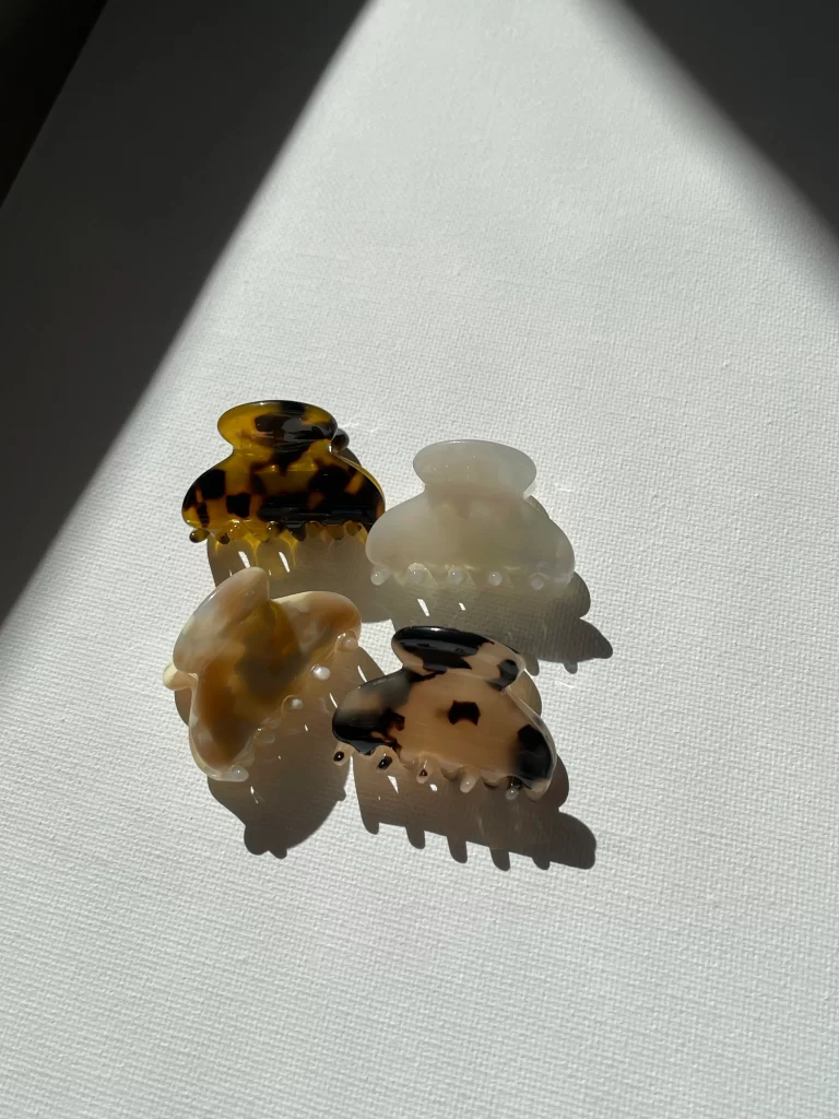 Four small PixieWoo claw clips are shown, with different colors visible: one in brown and black, one in white, one in beige and white, and one in beige and black.